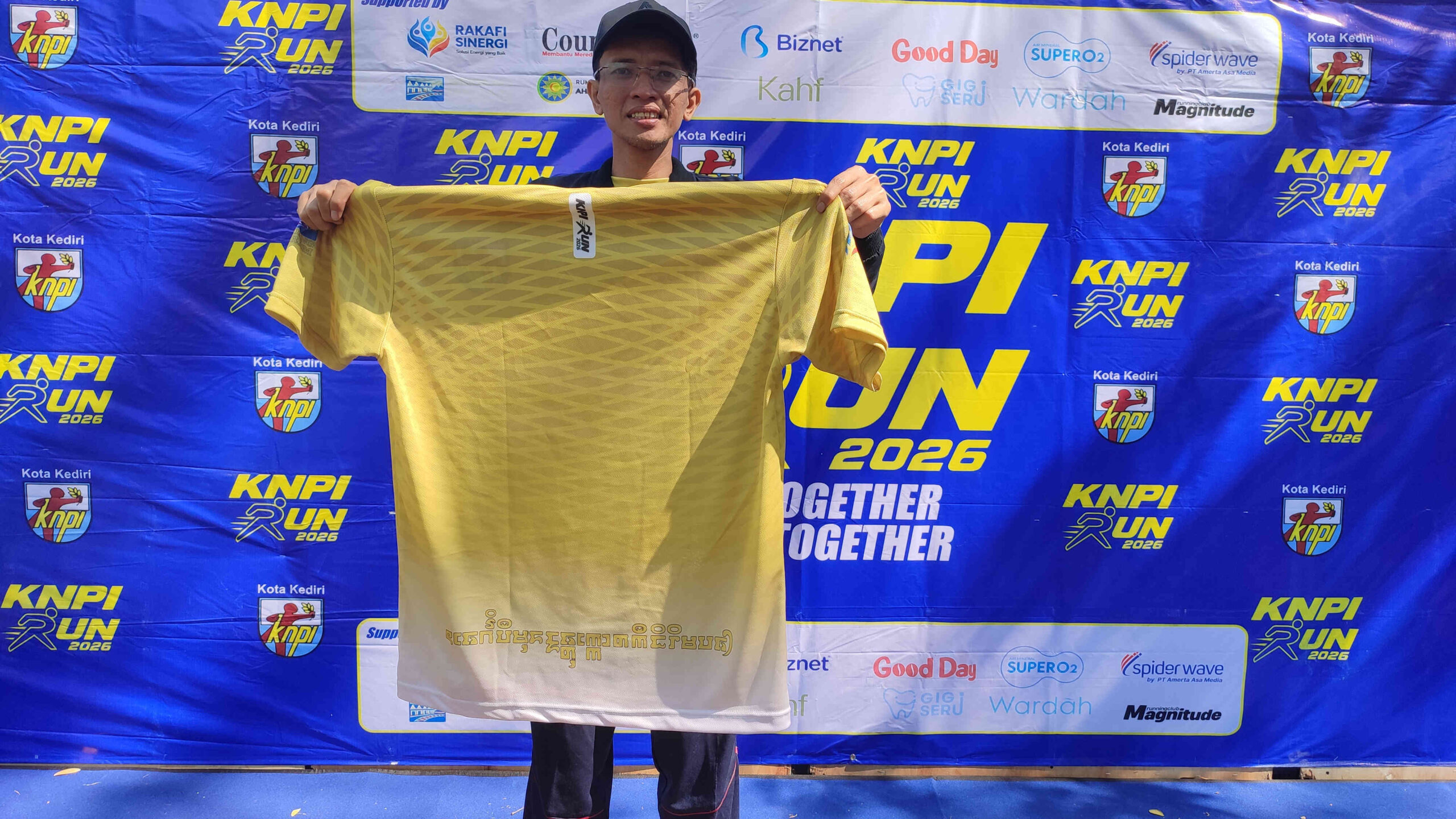 Ketua DPD KNPI Kota Kediri, Munjidul Ibad, memperlihatkan bagian belakang kaos resmi KNPI Run 2026 yang memuat tulisan Aksara Kawi Kwadrat "Energi Pemuda untuk Kota Kediri Mapan". Penggunaan aksara kuno tersebut menjadi bagian dari upaya KNPI Kota Kediri melestarikan budaya lokal sekaligus mengenalkan sejarah Kediri kepada generasi muda dalam rangka Harlah ke-53 KNPI, Minggu (26/7/2026).